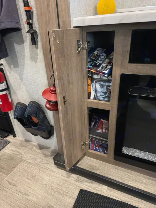 RV rental interior: Open cabinet with DVDs/Blu-rays (Westworld, Batman), fireplace, hanging shoes, and a lantern for entertainment and storage.. Grand Design Imagine 2023
