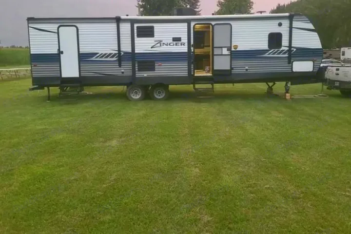This Zinger travel trailer RV rental is parked on a grassy campsite with its main door open and stabilizers extended.. Crossroads RV zinger 2019