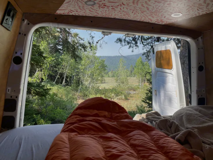 Inside an RV rental, a comfy sleeping bag faces open van doors revealing a lush forest and distant mountains.. Mercedes-Benz Sprinter 2018