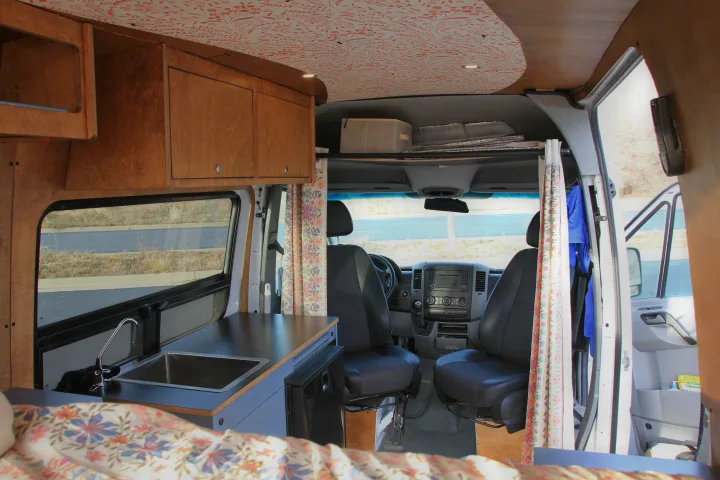 View inside an RV rental showing a wood-paneled kitchen with sink, built-in fridge, floral bed, and front driver's cab.. Mercedes-Benz Sprinter 2018