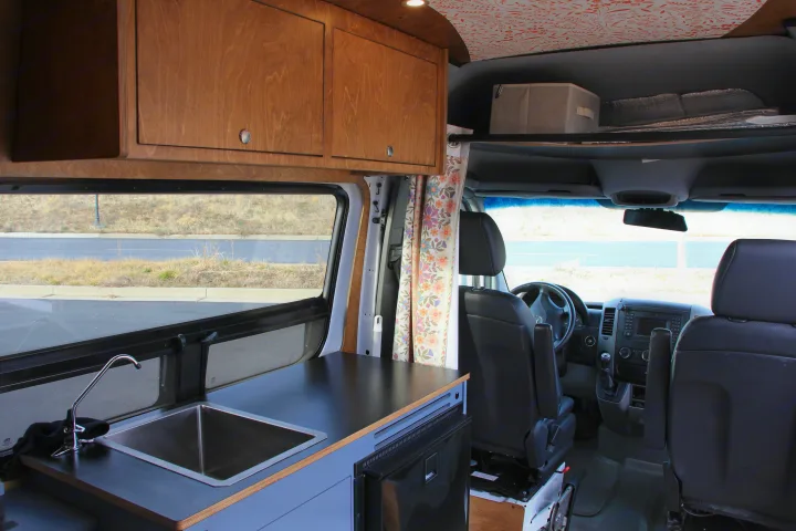 This RV rental features a camper van interior with a kitchenette, wooden overhead cabinets, and a view into the driver's cabin.. Mercedes-Benz Sprinter 2018