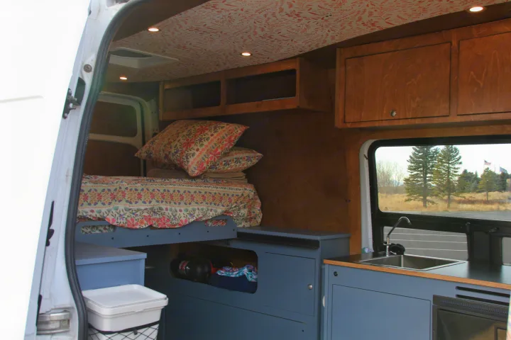 This rv rental shows a cozy, customized interior with a floral bed, blue storage, kitchen, and wood cabinets.. Mercedes-Benz Sprinter 2018