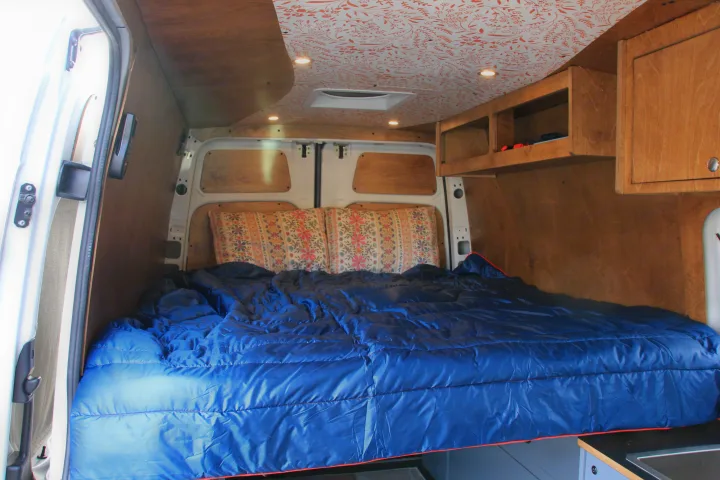 Inside an RV rental: Custom campervan interior featuring a bed with blue bedding, wood walls, cabinets, and a patterned ceiling.. Mercedes-Benz Sprinter 2018