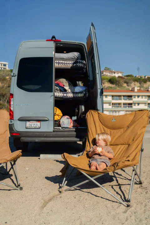 Toddler enjoys an apple in a beach chair beside a grey Sprinter rv rental with open doors, ready for family coastal adventure.. Mercedes sprinter 2025