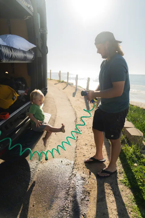 A dad rinses his child's feet by their camper van at the beach. Perfect for an rv rental family adventure by the ocean.. Mercedes sprinter 2025