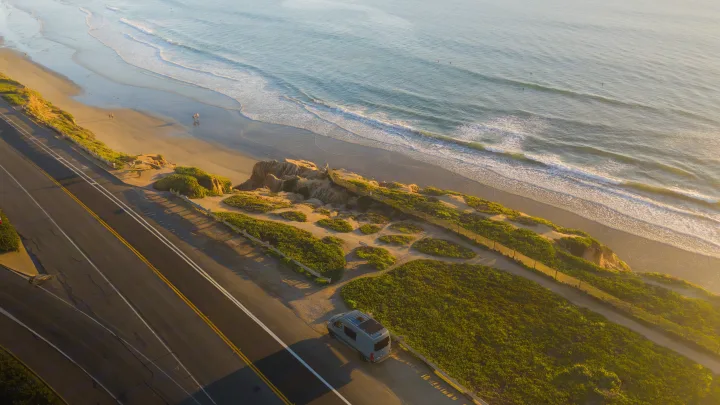 Coastal RV rental: A camper van parked by the road, overlooking a sunlit beach with waves, surfers, and distant people.. Mercedes sprinter 2025