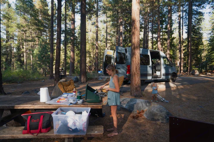 Family camping trip with an RV rental. A woman cooks at a picnic table; a child plays near their camper van in a forest.. Mercedes sprinter 2025