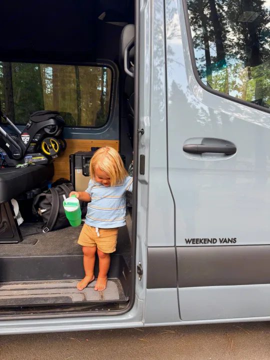 A blonde toddler in a "Weekend Vans" RV rental, holding a green cup in the doorway, enjoying nature.. Mercedes sprinter 2025
