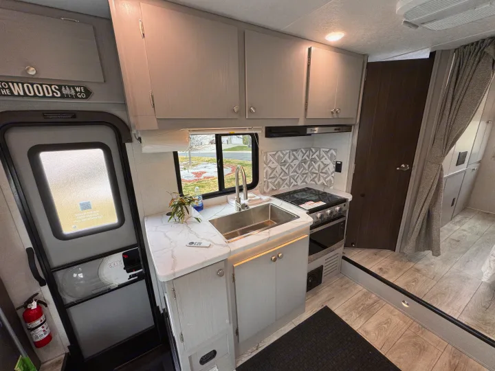 Spacious RV rental kitchen with modern cabinets, sink, stove, and a welcoming "To the Woods" sign by the entry door.. Forest River Coachmen Freelander 2026