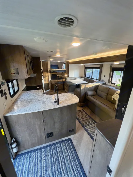 Modern RV rental interior with kitchen island, dinette, sofa, and bunk beds. Features light wood tones & blue rugs.. Forest River Wildwood X-Lite 2020