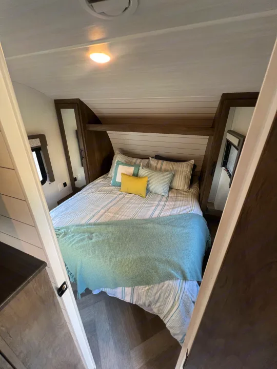 Modern RV rental bedroom features a cozy bed with striped linens, blue throw, built-in shelf, and windows. Ideal for travel.. Forest River Wildwood X-Lite 2020