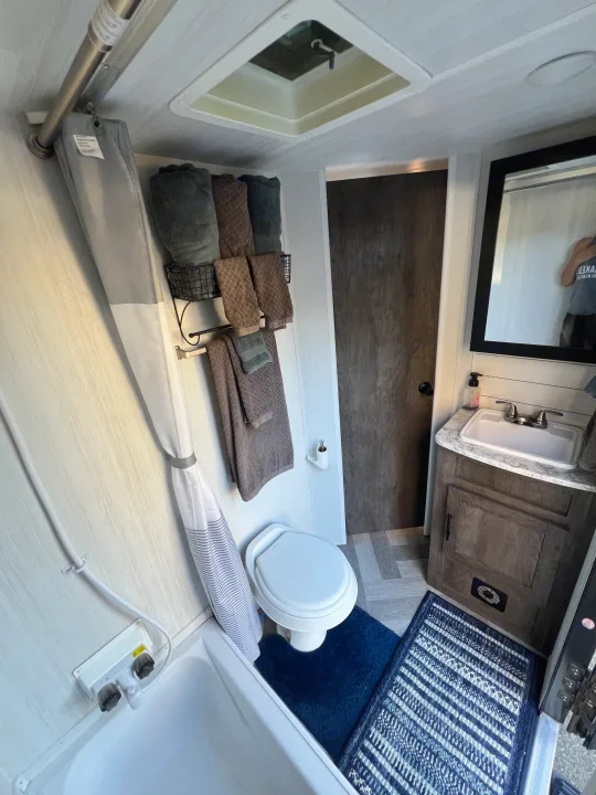 A compact RV rental bathroom featuring a shower, toilet, sink vanity, and neatly organized towels. White walls, wood accents.. Forest River Wildwood X-Lite 2020