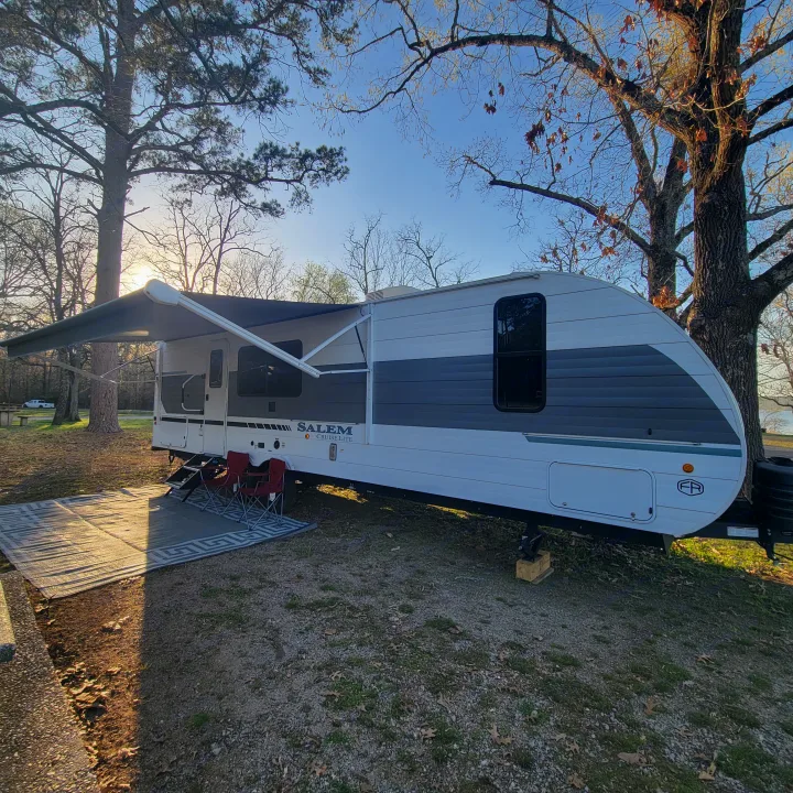 This comfortable rv rental travel trailer is set up at a lakeside campsite with an awning, chairs, and mat.. Forest River Salem Cruise-Lite 2026