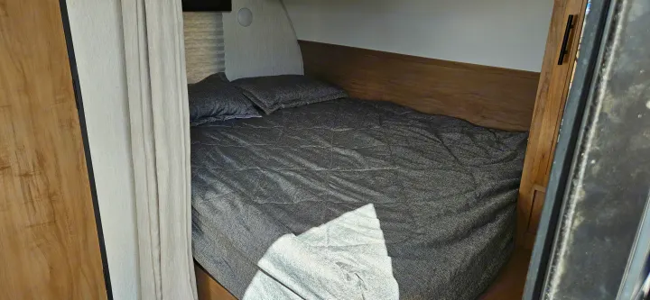 Compact RV rental bedroom with gray quilted bed and pillows, wooden headboard, and curtains, offering a cozy sleeping area.. Forest River Cherokee Grey Wolf 2023