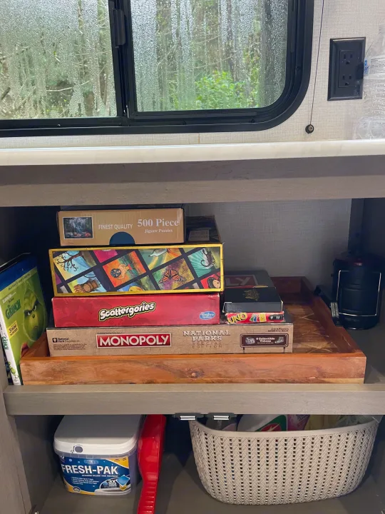 Board games, puzzles, and RV supplies are neatly stored in an RV rental cabinet, with a rainy window showing trees.. Grand Design Transcend Xplor 2026