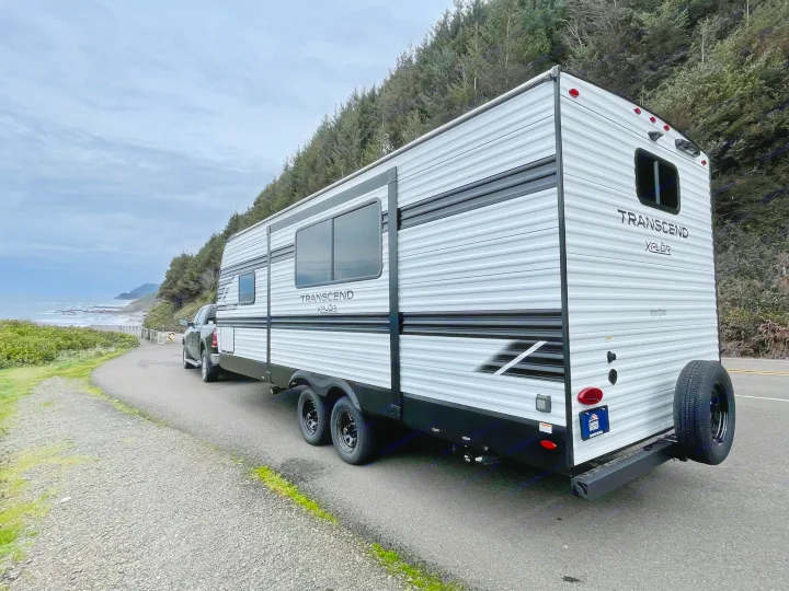 An RV rental, a white Transcend Xplor travel trailer, parked by a scenic coastal road with ocean views and forested hills.. Grand Design Transcend Xplor 2026