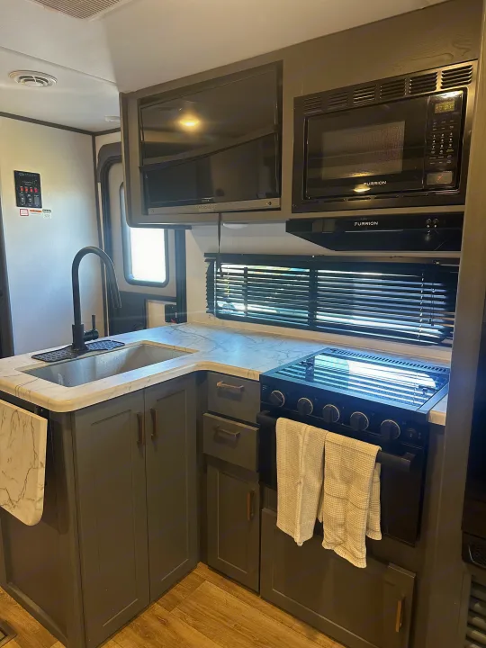 Modern RV rental kitchen showing a sink, stove, microwave, and TV. Features include gray cabinetry and a light marble-look countertop.. Keystone RV Bullet Crossfire 2024