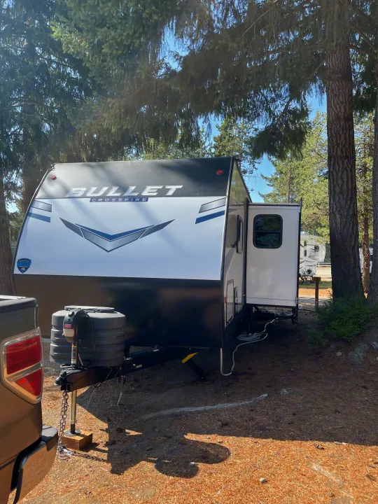 A Bullet Crossfire travel trailer RV rental, featuring white and black exterior, parked at a wooded campsite with tall pines.. Keystone RV Bullet Crossfire 2024