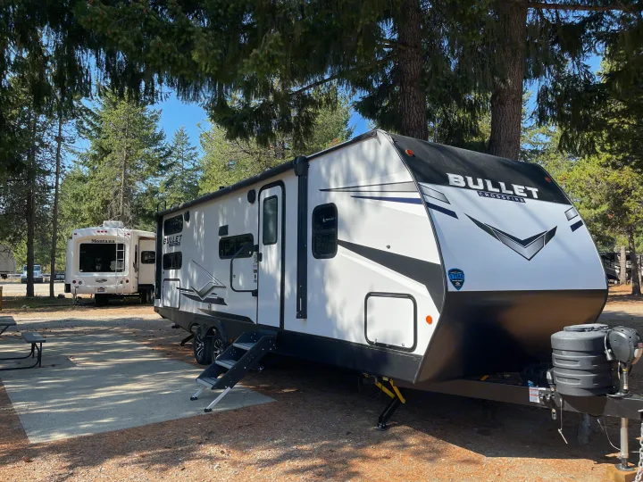 A white Bullet Crossfire travel trailer, a great rv rental option, parked at a shaded, wooded campsite with pine trees.. Keystone RV Bullet Crossfire 2024
