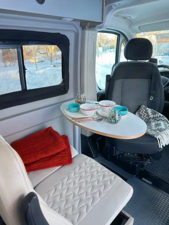 Cozy RV rental interior with a dining setup for two, featuring a swivel table, bench seat, and snowy views outside.. Winnebago Solis 2022