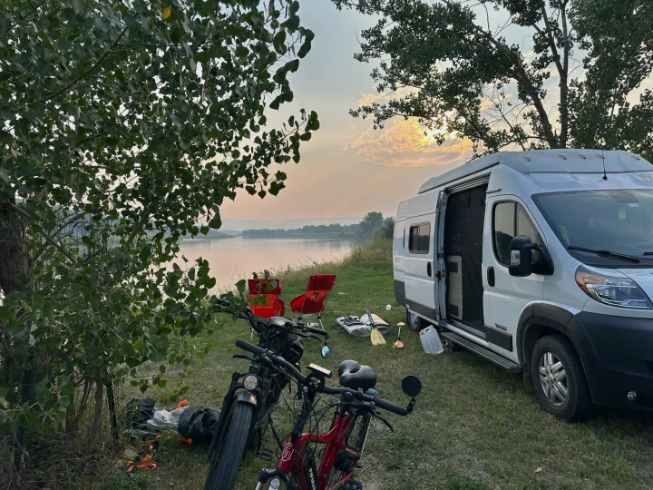 RV rental parked riverside at sunset. Bikes and camp chairs await next adventure by the calm water & trees.. Winnebago Solis 2022