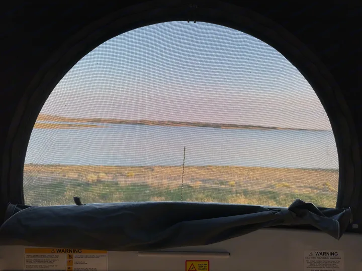 Inside an RV rental: Screened window frames a scenic lake and grassy landscape. Warning labels visible below.. Winnebago Solis 2022