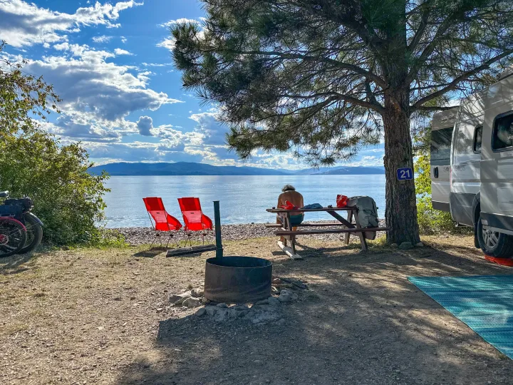 RV rental parked at site 21, a scenic lakefront campsite. Person sits at picnic table, red chairs on shore, fire pit nearby.. Winnebago Solis 2022