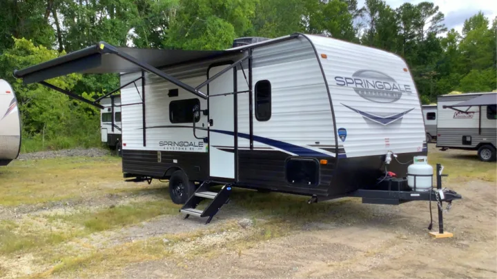 A Springdale Keystone travel trailer, ideal for an RV rental, with extended black awning and steps on a gravel lot.. Keystone RV Springdale 2023