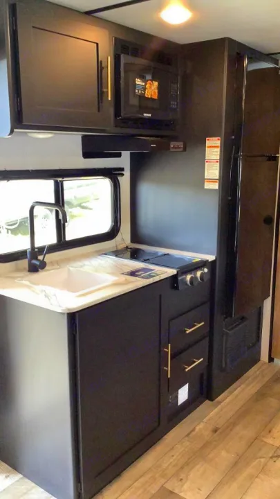 This RV rental's modern kitchenette features dark cabinets, a white sink, two-burner stove, microwave, and refrigerator.. Keystone RV Springdale 2023