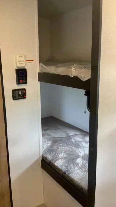RV rental interior featuring stacked bunk beds; the top bunk mattress is still plastic-wrapped. Control panels are on the wall.. Keystone RV Springdale 2023