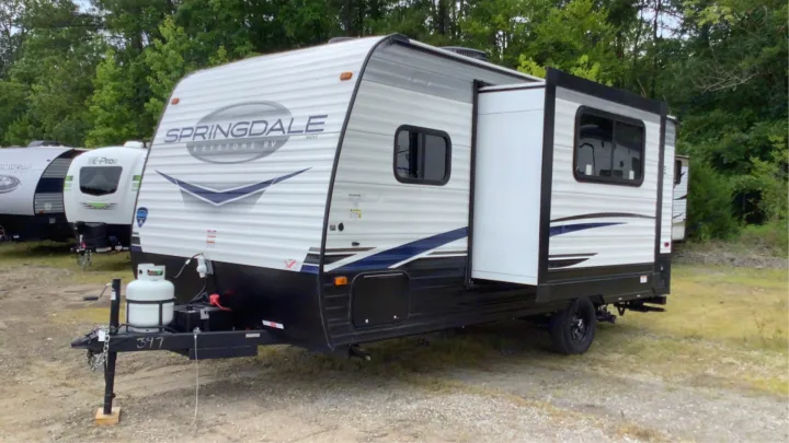 A white Springdale Keystone travel trailer RV rental with blue accents, parked on a gravel lot with trees.. Keystone RV Springdale 2023