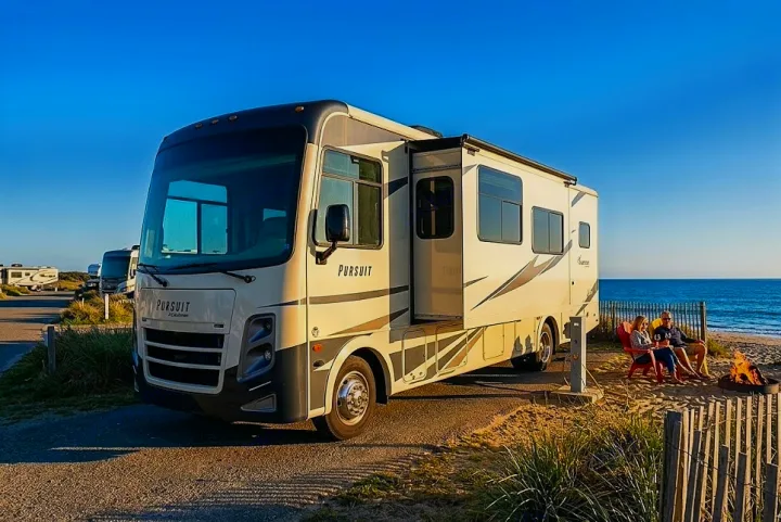 An RV rental is parked seaside, where a couple enjoys a beach campfire next to their motorhome with the ocean visible.. Coachmen Pursuit 2019