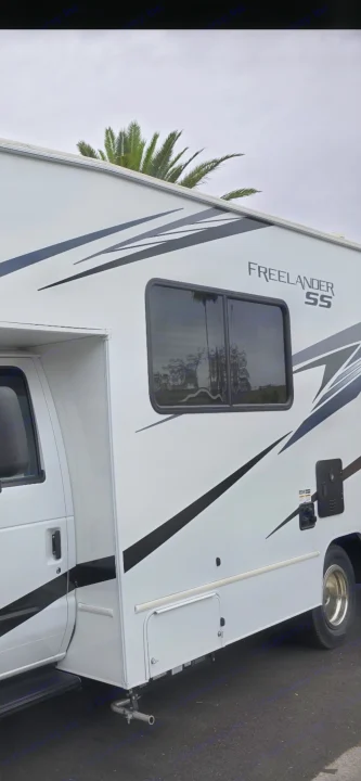 Side view of a white Freelander SS RV rental with black graphics, parked on asphalt. A palm tree is visible above.. Freelander 21QSS 2025