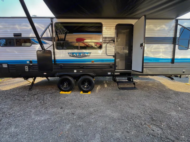 Modern RV rental travel trailer with open door, steps, and awning. Parked on gravel with leveling blocks under tires.. Forest River Salem 2023