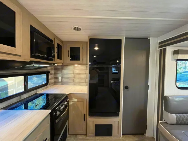 This modern RV rental kitchen features a sleek refrigerator, microwave, stove, and light wood cabinetry, with an adjacent seating area.. Forest River Salem 2023