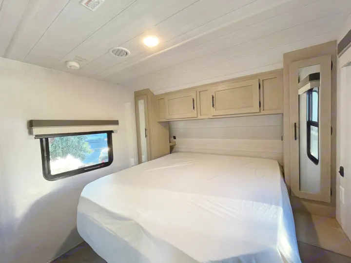 Bright RV rental bedroom features a bed with white sheets, overhead cabinets, and a window, offering comfort for travel.. Forest River Salem 2023