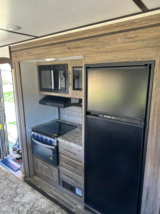 Compact RV rental kitchen featuring a modern microwave, cooktop, oven, and refrigerator in a wood-grain interior.. Crossroads RV Zinger 2021