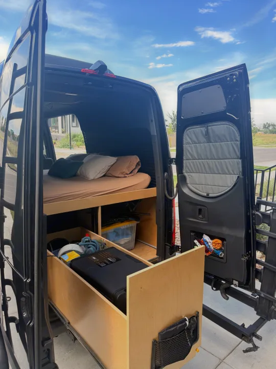 Rear view of an RV rental van's interior, showcasing a bed with pillows and an accessible pull-out storage drawer system.. Mercedez Benz Sprinter 2023