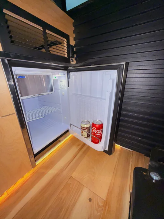 This RV rental shows an open Isotherm mini-fridge with drinks, built into a compact wooden interior. Perfect for road trip snacks.. Mercedez Benz Sprinter 2023