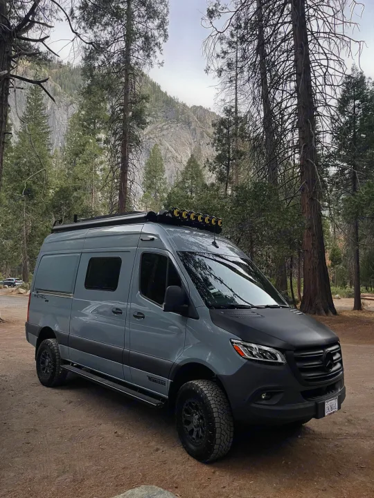 Adventure-ready gray Winnebago Revel RV rental in a mountain forest. Tall pines frame the customized van with roof lights & off-road tires.. Winnebago Revel 2022