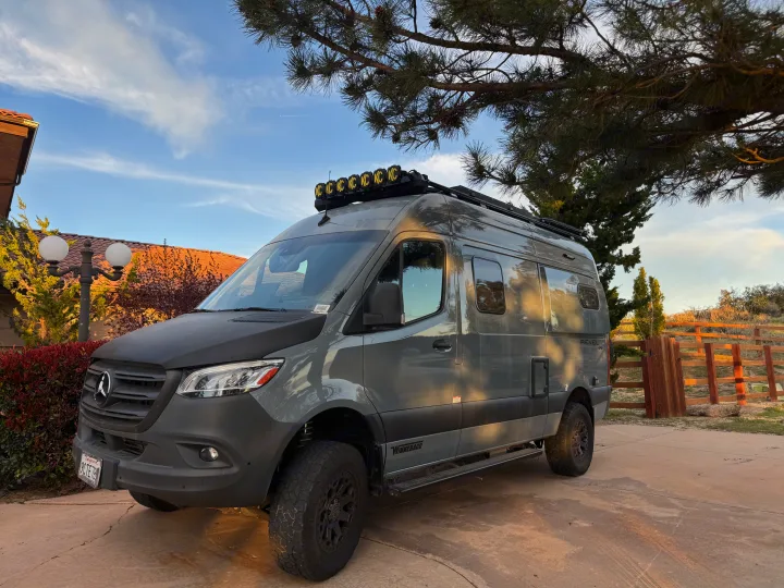 A sleek grey Mercedes Sprinter campervan, a Winnebago Revel 4x4, with off-road tires under a pine tree. Ready for your rv rental adventure!. Winnebago Revel 2022