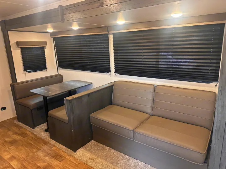 RV rental interior: a cozy dinette with light taupe booth seating and table, beside a matching sofa. Dark pleated blinds cover the windows.. Keystone RV Hideout 2022