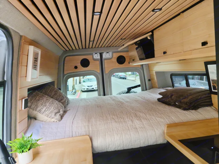 Cozy RV rental interior with a comfortable bed, wooden ceiling, and street view. Perfect for travel!. Mercedes-Benz Sprinter 2021
