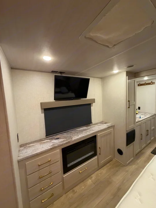 Modern rv rental interior featuring a TV, electric fireplace, built-in storage, and a partial view of a bathroom vanity.. Forest River Surveyor Land Surveyor 2024
