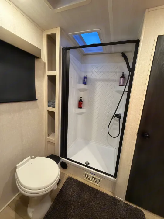Compact RV rental bathroom with a white toilet, enclosed shower with black frame, integrated shelving, and a bright skylight above.. Forest River Surveyor Land Surveyor 2024