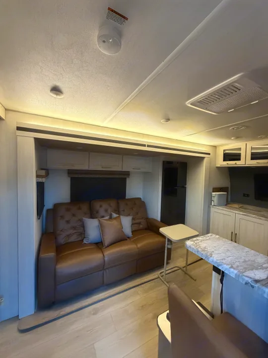Cozy RV rental living area with a brown leather sofa, light wood flooring, and modern kitchen counter under bright lighting.. Forest River Surveyor Land Surveyor 2024