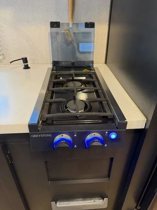 Greystone 2-burner gas stovetop in an RV rental kitchen, featuring bright blue illuminated controls and an open glass lid.. Forest River Cherokee Wolf Pup Black Label 2021