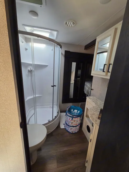This RV rental bathroom offers a compact corner shower, toilet, and vanity, with a laundry basket on the wood floor.. Starcraft Telluride 2021