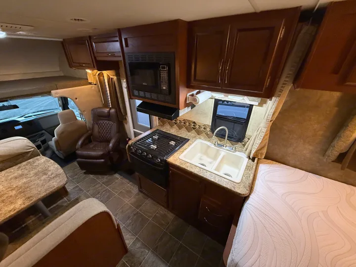 Cozy RV rental interior: kitchen with stove, sink, microwave, dining area, armchair, and bed. Features wood cabinetry & tiled floor.. Thor Freedom Elite 2012