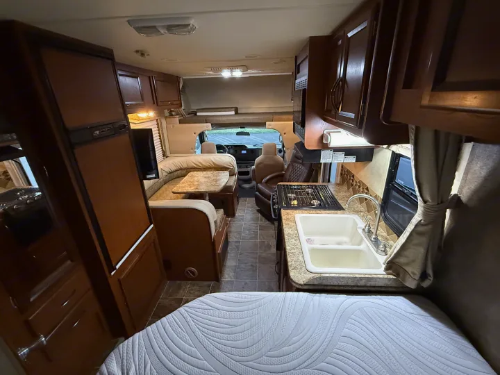 Inside this cozy RV rental: kitchen with sink & stove, dining booth, refrigerator, and a bed in the foreground.. Thor Freedom Elite 2012